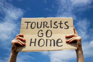 Metin Turistler ellerinde bir karton plaka üzerinde eve gidin. Bulutların arka planı olan mavi gökyüzü. Kitle turizmi kavramını durdurun.
