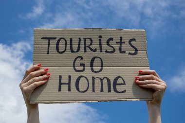 Metin Turistler ellerinde bir karton plaka üzerinde eve gidin. Bulutların arka planı olan mavi gökyüzü. Kitle turizmi kavramını durdurun.