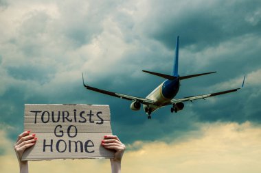 Bir insan bir uçağı ellerinde ki karton parçasıyla eve giden turistlerin elindeki bir kartonla selamlıyor. İnsanın arkasında bulutlar olan mavi bir gökyüzünde ki uçak. Kitle turizmi kavramını durdurun.