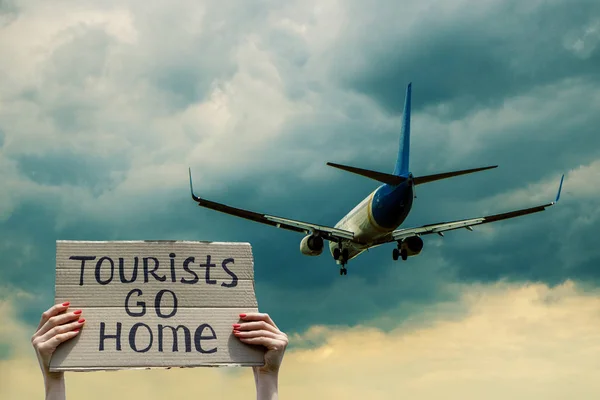A human greets a plane with text Tourists go home on a piece of cardboard in hands. Plane in a blue sky with clouds behind human. Stop mass tourism concept.