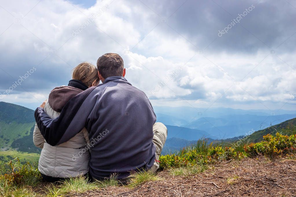 Happy couple of tourists during a travel in mountains, stand on the top and looking at the cloudy sky and the valley.