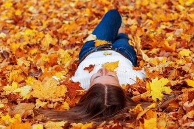 Yetişkin bir kadın küçük bir kız gibi sorunsuz hissediyor. Kot pantolonlu ve beyaz kazaklı kadın, düşen sarı kuru sonbahar yapraklarının üzerinde yatıyor. Ön planda kal. Parkta ve ormanda dinlen..