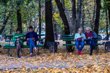 Lviv, Ukrayna - Ekim 2019. Parktaki banklarda oturan iki çift: gençler kitap okuyor, yaşlılar akıllı telefon kullanıyor. Kitaplar akıllı telefona karşı, aileler çocuklara karşı konsepti.