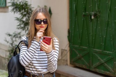 Sokakta çizgili gömlek ve güneş gözlüğü takan güzel bir kadının portresi. Şüpheci bir ifadesi olan kadın akıllı telefondan bir mesaj okuyor..