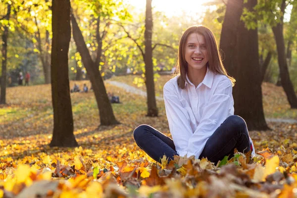 Sonbahar parkında düşen yaprağın üzerinde oturan gülümseyen genç kadın. Gün batımında arka plan ışığı.