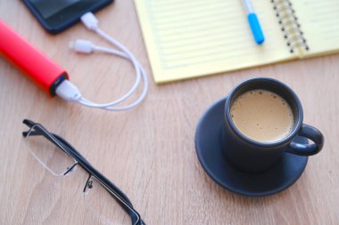 Akıllı telefon poverbank 'tan şarj oluyor. Masada yakın çekim kahve, günlük, kalem ve gözlük bir fincan.