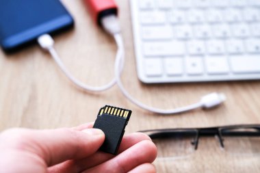 Hafıza kartı eldeki. Akıllı telefon güç kaynağından şarj edilir. Masada yakın çekim kahve, klavye ve gözlük bir fincan.