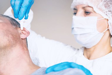 Doktor Otolaryngologist otitis media hasta muamelesi yapıyor. Kulak damlası kulak deliğe gömer. Koruyucu maske yüzünde. Koruyucu elbise.