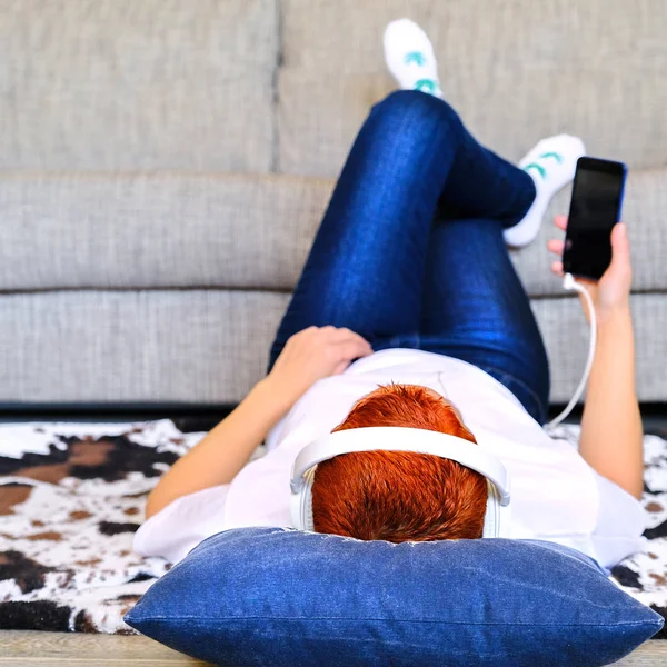 Een leuk meisje in een koptelefoon gebruikt een gadget. Liggend op een deken. In de buurt van de Bank thuis. — Stockfoto