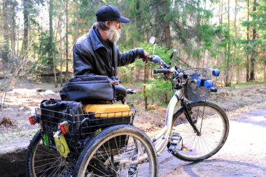 Homeless in a wheelchair going on a forest road. The three-wheeled wheelchair is equipped with a box for things. An unwashed bearded man. clipart