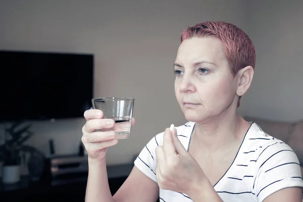 Dolor desagradable. Triste mujer hermosa triste sentada a la mesa. Depresión y dolor de cabeza. Toma una pastilla y se lava con agua . — Foto de Stock