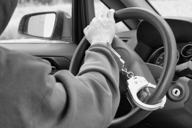 Siyah beyaz fotoğraf. Hava korsanı direksiyona kelepçelenmiş. Suçlu bir araba çalarken yakalandı. Polis iyi iş çıkardı.