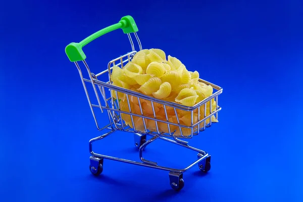 Pasta in de vorm van schelpen, mini supermarktkarretje naar boven gevuld. Ontwerp voor elk doel van reclame producten. Blauwe achtergrond. — Stockfoto