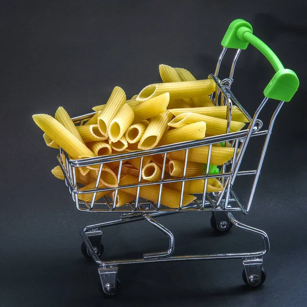 Pasta in de vorm van gesneden buisjes, een mini supermarktkarretje tot aan de rand gevuld. Donkere achtergrond. Ontwerp voor reclamedoeleinden. — Stockfoto