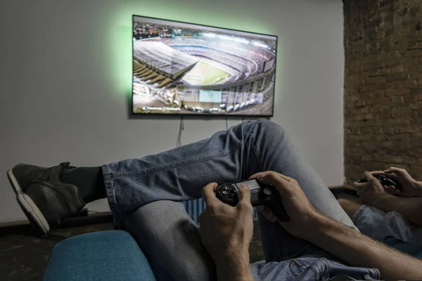 Two men playing video game. Hands holding console controller. Football or soccer game on the television. Widescreen tv hang on the wall