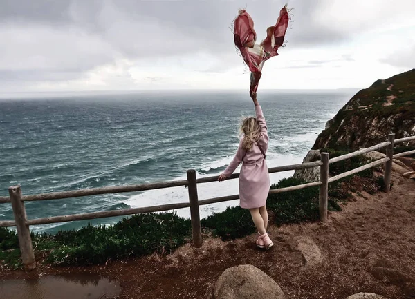 A blonde in a pink raincoat is standing outdoors at the edge of a cliff at Cape Roca in Portugal and holding a scarf in the wind. The concept of happiness and freedom