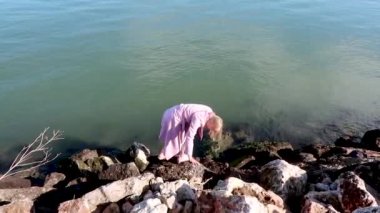 Pembe paltolu ve botlu genç sarışın kadın güneşli havada büyük deniz taşlarının üzerinde yürür, suya düşmemeye çalışır. Kaygan taşlar, rahatsız edici kıyafetler..