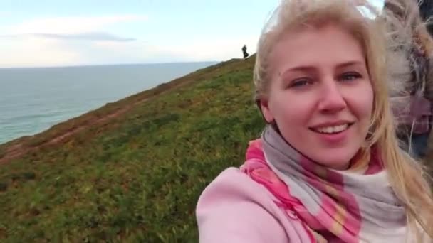 ビデオの若いブロンドの女性は 美しい風景の背景で示す海のスマート フォン チャットします 冬で崖の上笑っているエキサイティングな女の子 強い風に舞う髪 ポータブル ハンドヘルド デバイスを使用しています Selfie — ストック動画