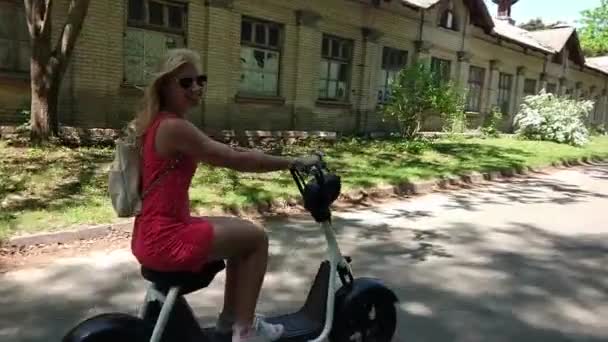 穿着红色礼服的年轻金发碧眼的女子在公园里骑电动摩托车时微笑着 — 图库视频影像