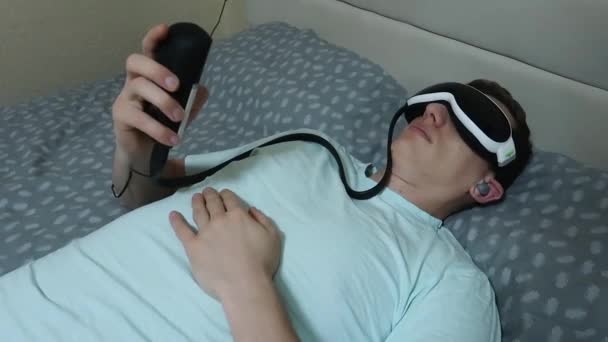 年轻迷人的男人使用眼压装置 躺在床上 拿着遥控器 舒缓疲劳 在笔记本电脑上工作后休息 — 图库视频影像