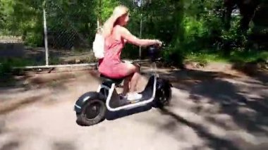 Sırt çantası ile kırmızı elbiseli sarışın yakışıklı genç kadın parkta elektrikli scooter üzerinde sürme sırasında gülümsüyor