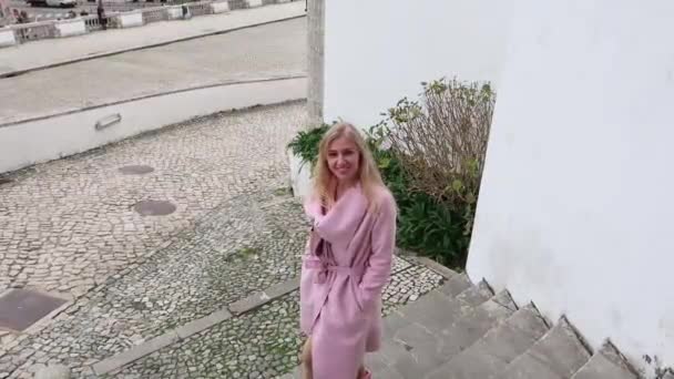 迷人的金发碧眼的女人 穿着粉红色的外套 在楼梯上慢慢地下来 看着镜头 微笑着 — 图库视频影像