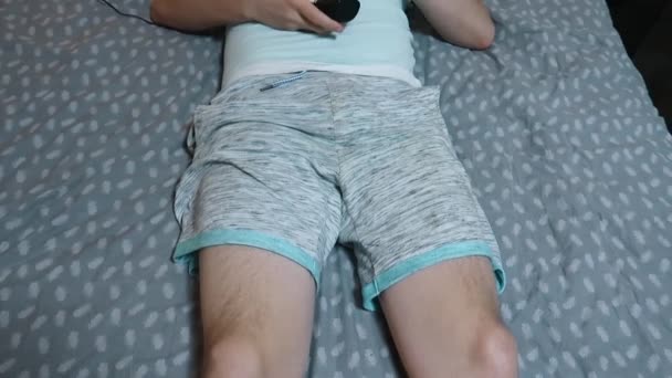 若い魅力的な男性は 目のマッサージ装置を使用して ベッドの上に横たわって リモートコントロールを保持し 疲労を和らげ リラクゼーション ノートパソコンで働いた後の休息 上からの眺め — ストック動画