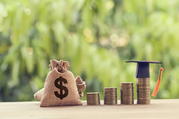 Siyah mezuniyet şapkası, bir masanın üstünde yükselen bozuk paraların üzerinde şapka ve Amerikan Doları çantası. Eğitim fonu, finansal konsept. İleriki çalışmalar için çocuk bilgisi birikimlerini gösterir