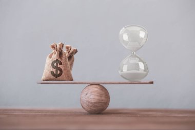 Kum saati ya da kum saati sembolü ve dolar çantasına eşit bir denge terazisi yazın. Finansal konsept: Paranın zaman değeri, zaman içinde varlık büyümesi, daha fazla para büyümesi için uzun vadeli sermayeye yapılan yatırımı resmediyor