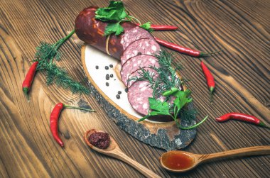 Sosis baharatlar ile dilimlenmiş, dereotu salata yaprakları, bir kesme tahtası üzerinde kahverengi ahşap masa arka plan üzerinde kırmızı biber biber ve sarımsak adet.