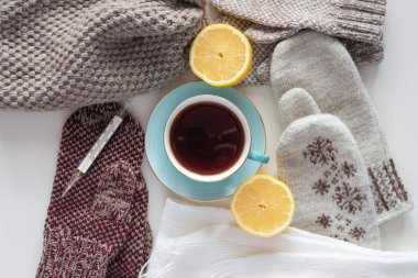 Bir fincan sıcak çay, termometre, limon ve eşarp beyaz masa arkasında..