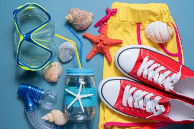 Yaz tatili konsepti düzdü. Plaj aksesuarları. Dalgıç maskesi, deniz kabukları, kırmızı denizyıldızı, şort, sakız ayakkabısı ve mavi arka planda kokteyl kavanozu..