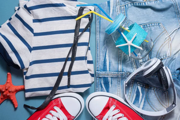 Red gum shoes, jeans shorts, shirt, red starfish, headphones and cocktail jar with tube on blue flat lay background. Summer vacation background.