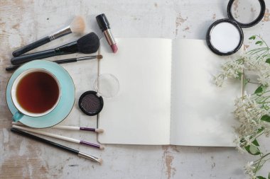Boş sayfa defterini aç, makyaj fırçaları, göz gölgeleri, bir fincan çay ve beyaz ahşap bir masada çiçek..