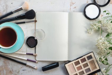 Boş sayfa defterini aç, makyaj fırçaları, göz gölgeleri, bir fincan çay ve beyaz ahşap bir masada çiçek..
