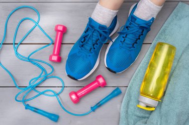 Spor ayakkabılı kadın bacakları, atlama ipi, havlu, halterler ve ahşap zemin üzerinde bir şişe su..
