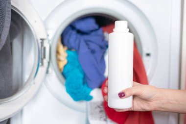 Kadın el ele çamaşır makinesinin arka planında sıvı çamaşır deterjanı tutuyor..