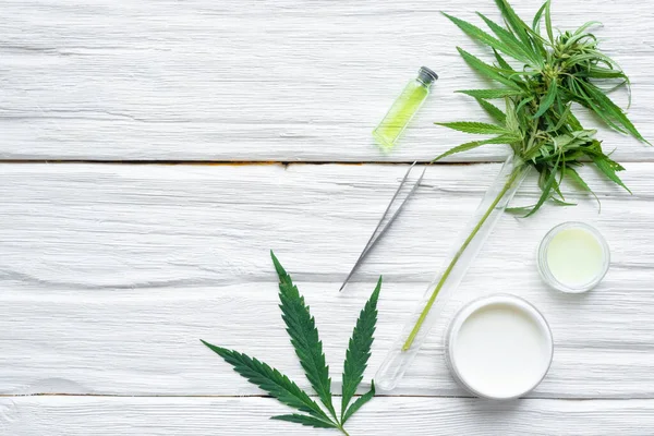 瓶の中の大麻の化粧品クリームとコピースペースのある白い木製のテーブルの背景の緑の植物の葉 天然化粧品 — ストック写真