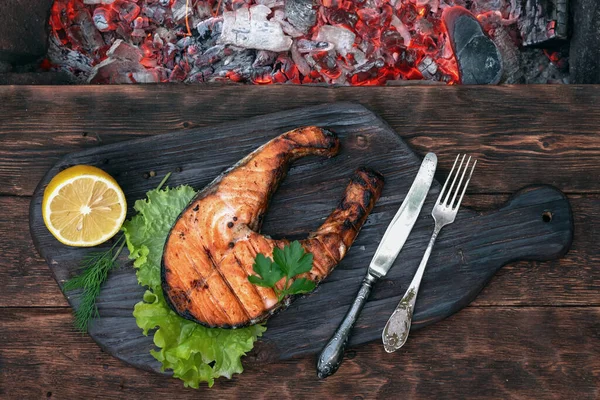 Grilled salmon steak on a old cutting board on a brown wooden table with a burning coals above. — Stock Image Grilled Salmon Steak Old Cutting Board Brown Wooden Table Burning Royalty Free Stock Photos