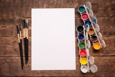 Fotokopi alanı, gouache kavanozları ve boya masası arka planında boya fırçaları olan boş beyaz sayfa.