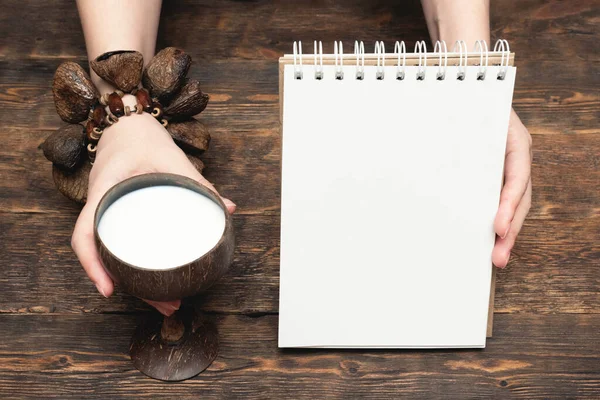 Coconut milk in cup and coconut and blank page notepad with copy space on brown wooden table background.