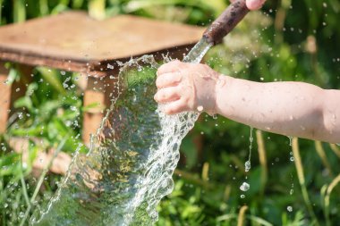 Küçük çocuk bahçedeki bir su hortumunun altında elini yıkıyor..