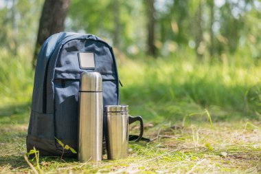 Sırt çantası, süpürge şişesi ve çelik bardak yeşil orman arka planında..