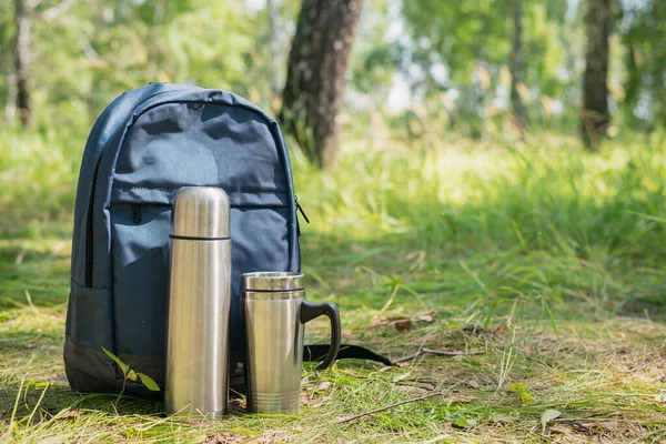 Tourist backpack, vacuum flask and steel cup on green forest background.