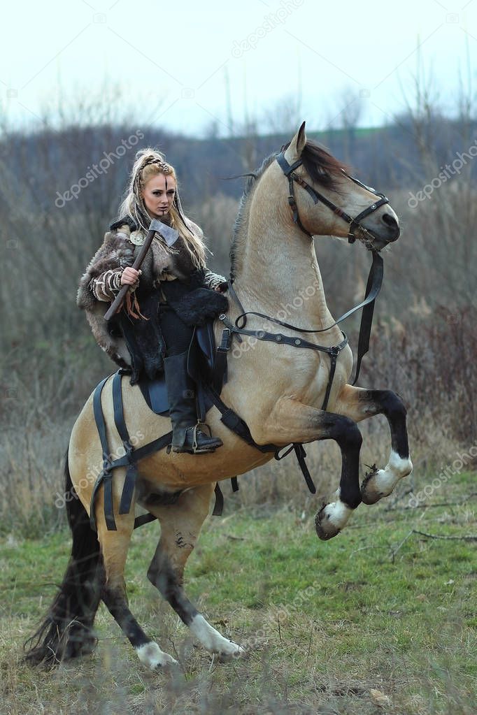Blonde Viking warrior woman riding a horse with ax in hand. Scandinavian warrior woman in forest with war makeup, ready to attack