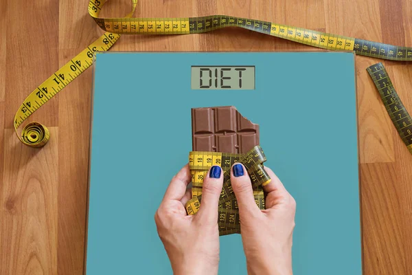 Hands of woman with on weighing scale asking for help to avoid the temptation to eat sweets. Stop eating sweets, healthy diet concept with measurement tape rolled on chocolate above weight scale