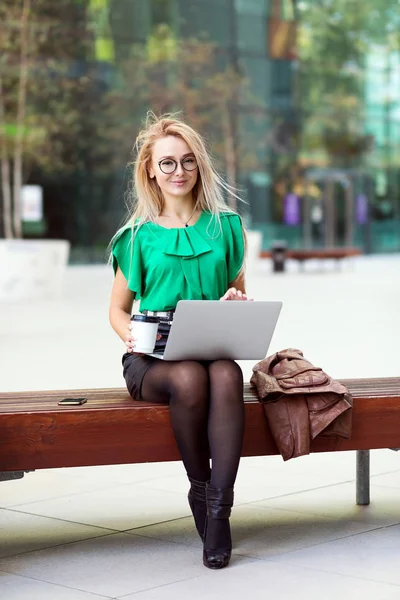 Young eastern European businesswoman walking on laptop on coffee break outside office building in windy day. Woman sitting on a bench holding a to go coffee while checking emails.