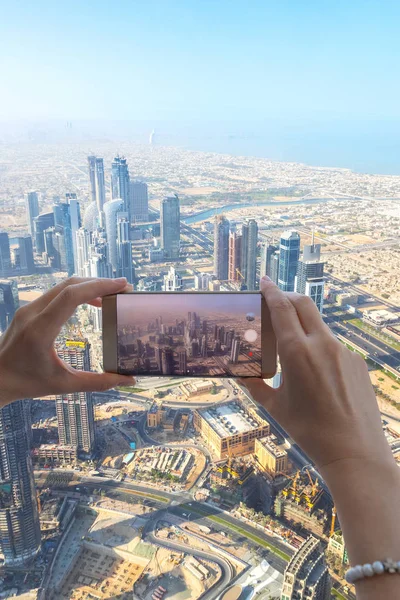 Closeup of woman tourist hand taking photo of Dubai city with smartphone camera from Burj Khalifa tower. Girl taking picture with mobile phone on holiday to post on social media