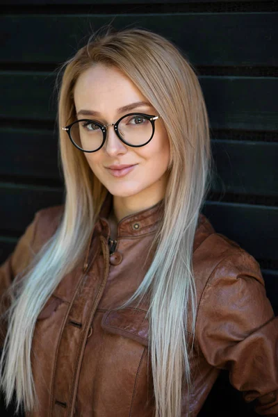 Beautiful young blonde woman with stylish glasses outdoor against dark background. Concept of young girl wearing glasses and seeing the world with new eyes
