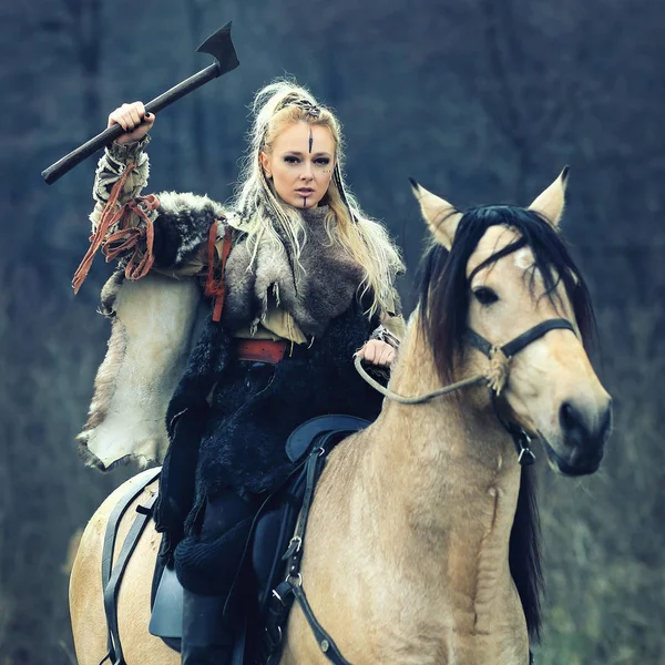 Beautiful viking warrior woman holding ax in hand wearing traditional warrior clothes riding a horse. Woman warrior with braided hair and makeup on horse in forest.